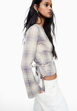 Mlada ženska, oblečena v oprijet top iz kariranega materiala z dolgimi rokavi in zavezovanjem na straneh ter bele hlače z visokim pasom, stoji v profilu na belem ozadju.