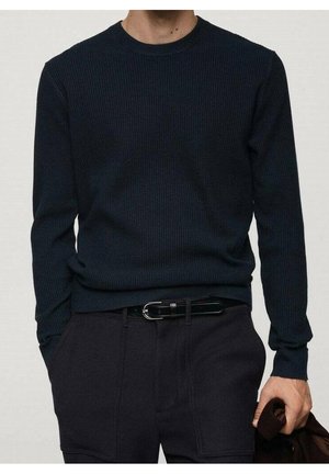 Homme portant un pull à manches longues côtelé bleu foncé et un pantalon noir avec une ceinture noire, tenant une veste sombre pliée dans une main.
