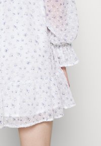 Vit blommig klänning med puffärmar, ett lager på lager volangkant och ett lätt, texturerat tyg med små blå och gröna blommönster.