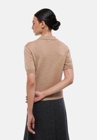 Pullover polo à manches courtes de couleur brun clair avec une texture côtelée, doté d'un col et d'une coupe ajustée, porté avec une jupe gris foncé.