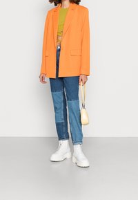 Osoba v oranžovém saku, zeleném krátkém topu, prošitých modrých džínách, bílých šněrovacích botách a s malou žlutou kabelkou.