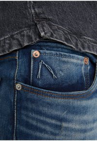 Siniset farkut, joissa haalistunut pinta, kupariset ja hopeiset niitit sekä takataskussa tikattu nuolikoriste.