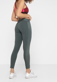 Leggings sportivi verde scuro a vestibilità slim, con tre righe verticali e una cintura larga. Abbinati a scarpe da ginnastica bianche.