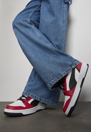 Personne portant un jean large bleu et des baskets Puma blanches, rouges et noires avec des lacets noirs, se tenant sur un sol gris contre un mur blanc.
