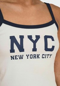 Top bianco senza maniche con bordi blu navy e grande testo stampato "NYC" e "NEW YORK CITY" in lettere in grassetto. Texture a coste.