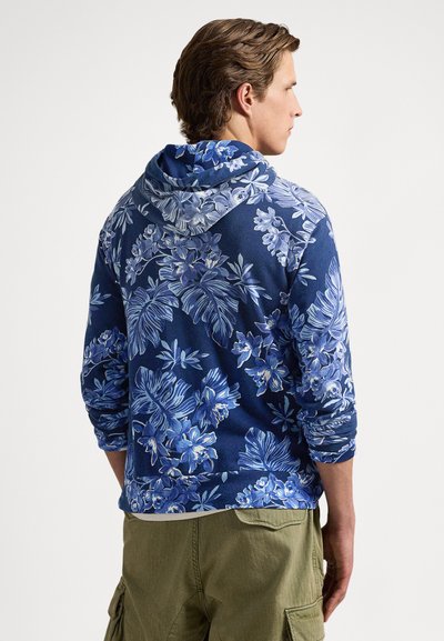 Homem vestindo um capuz azul com padrão floral, capuz abaixado, calças cargo verde-oliva, de costas contra um fundo simples.