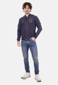 Giubbotto da aviatore navy con zip frontale, grande patch sul petto, due tasche laterali, polsini e orlo a coste, abbinato a jeans blu e sneaker chiare.