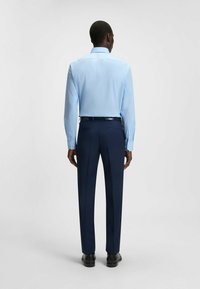 Camicia blu chiaro con bottoni, a maniche lunghe, abbinata a pantaloni eleganti di colore blu scuro, vista dal retro, che evidenzia il design elegante e la vestibilità.