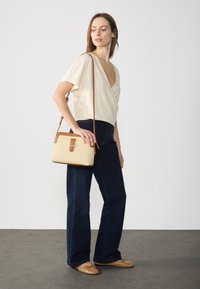 Donna in blusa avvolgente color crema, jeans blu scuro, ballerine color sabbia e che porta una borsa a tracolla intrecciata beige con rifiniture in pelle marrone.