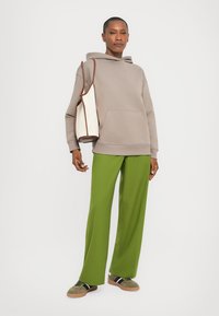 Haut à capuche gris avec poche, pantalon large vert, et baskets grises avec des rayures noires et blanches. Le mannequin porte un sac beige et marron.