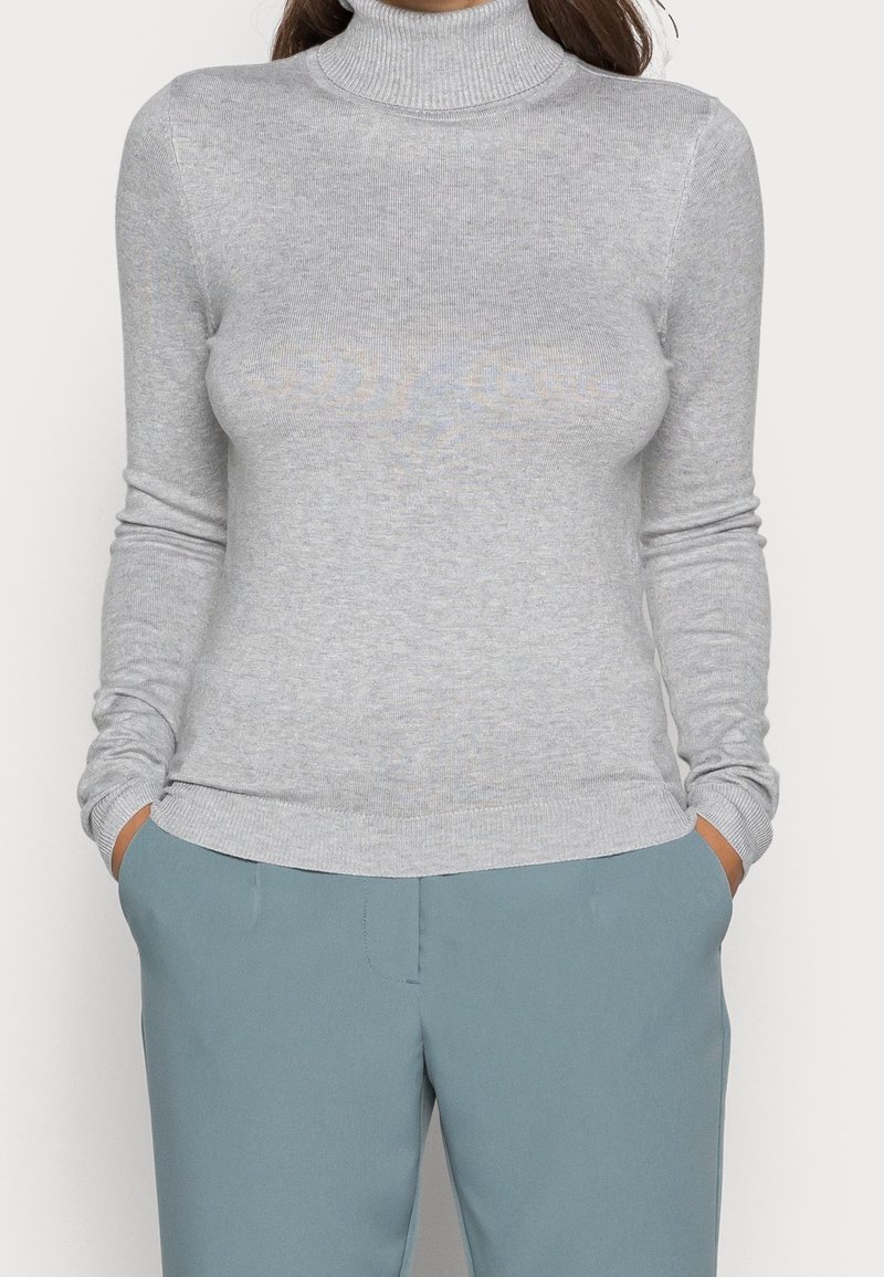 Personne portant un pull à col roulé côtelé gris clair et un pantalon bleu, les mains dans les poches, debout devant un fond uni.