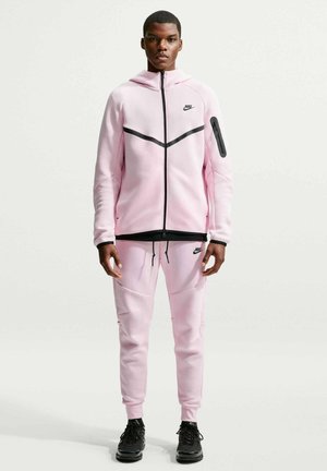 Mann steht und trägt einen hellrosa Nike-Trainingsanzug mit schwarzen Akzenten und schwarzen Sneakers vor einem schlichten weißen Hintergrund.