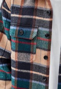 Poche de veste à carreaux boutonnée avec des boutons noirs dans des tons beige, vert, rouge, bleu et noir sur une chemise blanche.