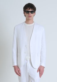 Blazer ajustado de lino blanco con cuello de chal, cierre de un solo botón y bolsillos de parche, combinado con un suéter ligero y pantalones a juego.