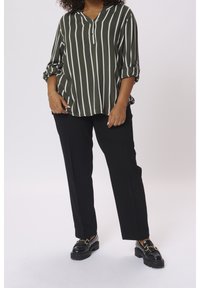 Chemise vert olive à rayures verticales blanches, manches retroussables et col en V. Associée à un pantalon noir et des mocassins noirs avec des accents dorés.