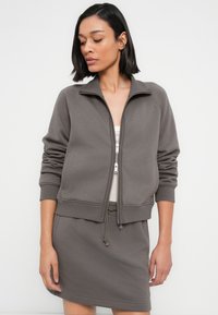 Γκρι ζακέτα με φερμουάρ, με ριμπ μανσέτες και κάτω μέρος, σετ με φούστα με κορδόνι στην ίδια απόχρωση. Μαλακό ύφασμα, απλός σχεδιασμός και ελάχιστες λεπτομέρειες.