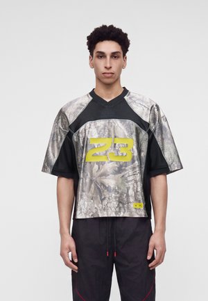 Jeune homme portant un maillot de sport à motif camouflage avec le numéro 23 en jaune, debout devant un fond clair uni.