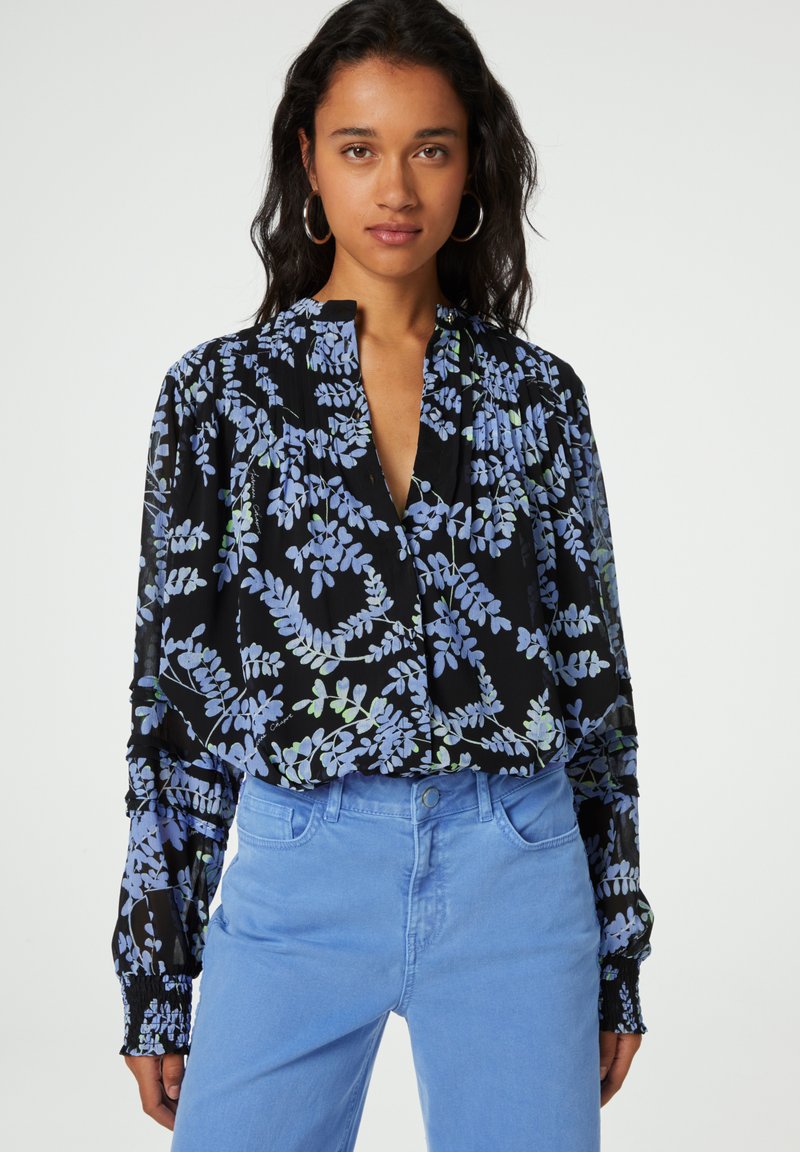 Schwarze Bluse mit blauem und grünem Blattmuster, tiefem V-Ausschnitt, gerafften Ärmeln und lockerer, langer Passform; kombiniert mit hellblauen Hosen.