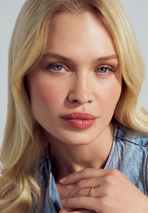 Tæt på en person med langt, blondt hår, blå øjne, og iført en denimskjorte. En delikat guldring med små accenter sidder på deres finger.