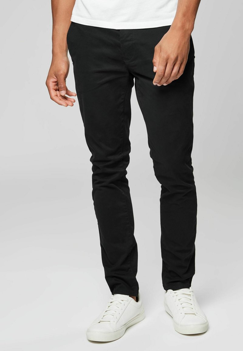 Črne slim-fit chinos hlače iz bombažne tkanine, z gladko teksturo in v kombinaciji z belimi športnimi copati. Jasno šivanje in minimalni oblikovni detajli.