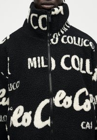 Μαύρο μπουφάν από φλις με λευκό μοτίβο κειμένου, ψηλό γιακά και μαύρο φερμουάρ στο μπροστινό μέρος, φορεμένο από ένα άτομο με σκουρόχρωμο δέρμα.