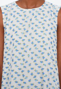 Haut blanc sans manches avec un motif de petites fleurs bleues et de feuilles vertes porté par une personne à la peau brune.