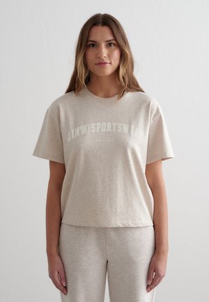 Kvinne som står iført beige matchende treningsantrekk med kortermet topp og bukser, ensfarget bakgrunn, avslappet holdning.