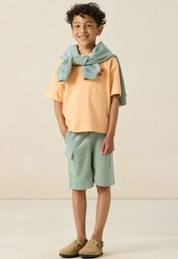 T-shirt orange clair avec texte rouge, shorts verts avec poches latérales, pull bleu clair drapé sur les épaules, et chaussures brunes à enfiler.
