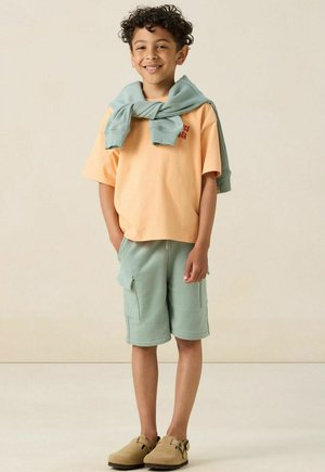 T-shirt orange clair avec texte rouge, shorts verts avec poches latérales, pull bleu clair drapé sur les épaules, et chaussures brunes à enfiler.