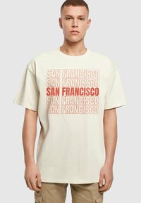 Béžové bavlnené tričko s opakujúcim sa textom "SAN FRANCISCO" v rôznych veľkostiach a štýloch, s voľným strihom a krátkymi rukávmi.