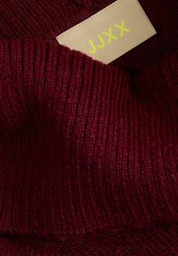 Vīna sarkanā džempera ar ribotu faktūru un dzeltenu zīmola etiķeti "JJXX". Redzamas šuves un mīksta auduma detaļas.