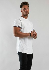 Vit t-shirt med rund hals, korta ärmar med mönstrad kant och en längre bakre fåll. Bärs med slim-fit svarta byxor.