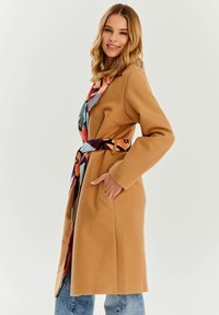 Płaszcz w kolorze wielbłądzim z długimi rękawami, z paskiem w talii i bocznymi kieszeniami. Posiada kolorową, wzorzystą podszewkę widoczną przy kołnierzu.
