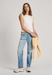 Top sin mangas blanco con detalles texturizados amarillos, jeans de mezclilla azul claro y un suéter de punto amarillo sostenido en la mano. Calzado informal completa el look.