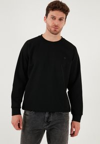 Črna trenirka iz bombaža, z okroglim izrezom in dolgimi raglan rokavi; na sprednji strani vsebuje droben logotip.