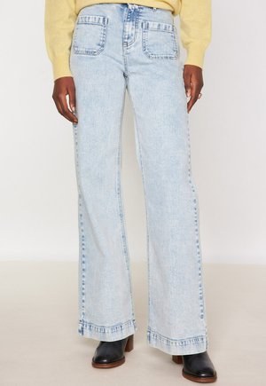 Model in een lichtblauwe hoge taille wijde jeans, gele trui en zwarte hoge hakken, staand tegen een witte achtergrond.
