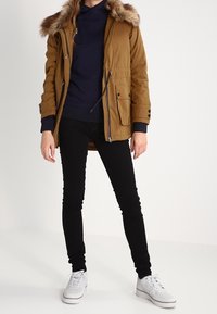 Brun parka-jacka med pälskantad huva, blixtlås och framfickor, kombinerad med en marinblå tröja och svarta tajta jeans, avslutad med vita sneakers.