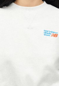 Κοντινή λήψη ατόμου που φοράει ανοιχτό γκρι φούτερ με μπλε κείμενο "new balance athletic shoes" και πορτοκαλί λογότυπο "NB" κεντημένο στο στήθος.