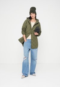 Πράσινο χιτώνα σε στυλ καπιτονέ με κουκούλα, μπεζ τοπ, σκισμένο ανοιχτό μπλε τζιν παντελόνι και μπεζ παπούτσια με μυτερή μύτη. Κρατά μια μικρή μαύρη τσάντα.