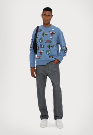 Blauwe sweatshirt met diverse patches, gecombineerd met grijze jeans en witte sneakers. Eenvoudig ontwerp met gestructureerde stof en geribde manchetten.