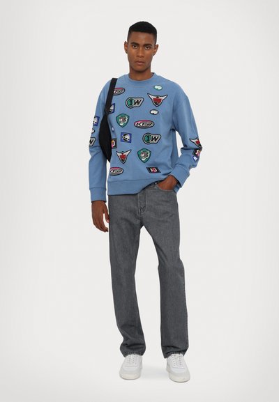 Felpa blu con vari patch, abbinata a jeans grigi e scarpe da ginnastica bianche. Design semplice con tessuto testurizzato e polsini a coste.