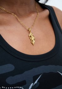 Colier cu pandantiv din aur, cu un design complex, prezentând modele spiralate. Atașat la o brățară din aur, purtat peste un top negru.