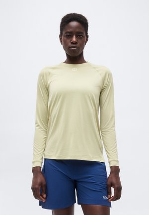 Vrouw in een licht beige shirt met lange mouwen en blauwe korte broek, staand met ontspannen armen tegen een effen witte achtergrond.