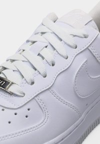 Biały skórzany sneaker z białymi sznurowadłami, perforowaną przednią częścią buta i zszytym logo Nike Swoosh z boku.