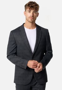 Dunkelgrau karierter Blazer mit einem einzigen Knopfverschluss, Reverskragen und drei-Knopf-Manschettendetails, über einem weißen T-Shirt getragen.