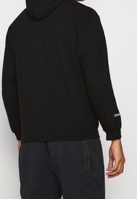 Sweatshirt à capuche noir avec des manches longues, des poignets et un ourlet côtelés, comportant un logo blanc sur la manche gauche. Tissu doux et texturé.