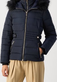 Doudoune bleu marine avec coutures horizontales, capuche doublée de fourrure et fermeture éclair frontale. Comprend des poches latérales et des accents métalliques.