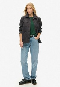 Vrouw in een zwarte jas over een groene shirt met lichtblauwe jeans en zwarte schoenen, staand met één hand in de zak op een witte achtergrond.