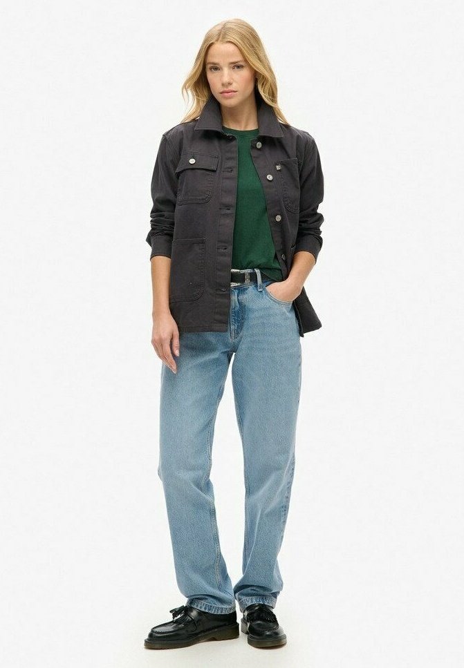 Vrouw in een zwarte jas over een groene shirt met lichtblauwe jeans en zwarte schoenen, staand met één hand in de zak op een witte achtergrond.
