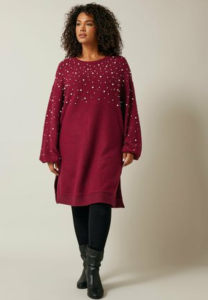 Robe longue bordeaux à manches longues, ornée de perles blanches sur la poitrine, avec des fentes latérales et un ourlet côtelé. Associée à des leggings noirs et des bottes.